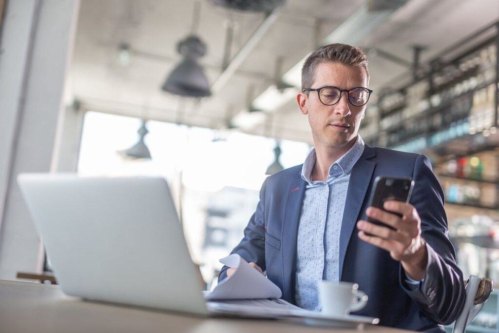 Young businessman types on his mobile phone with a laptop and coffee in front of him in a coffee shop.