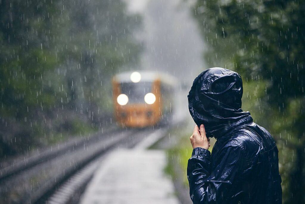 Man in drenched jacket standing on platform of railway station against train in heavy rain.